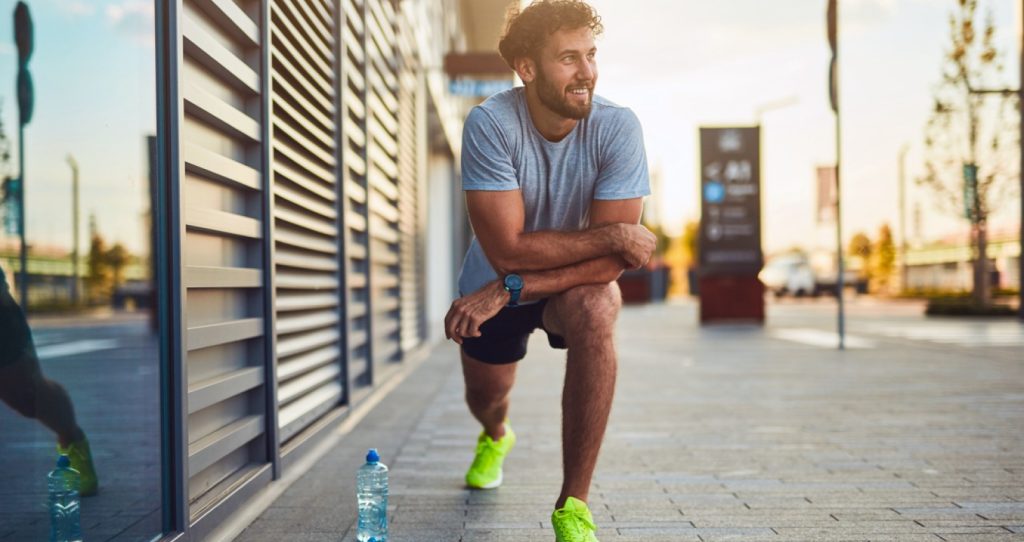 man with beard doing a lunge stretch on the side of the street and a bottle of water on the ground