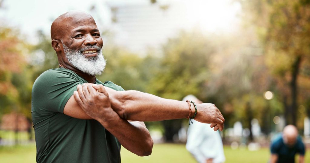 older man stretching his arm in the park while smiling and 2 other men in the background